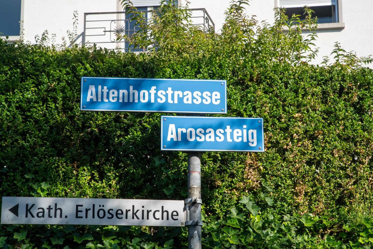 A metallic pole in front of a green hedge with signs to Altenhofstasse and Arosasteig, as well as an arrow sign to Kath. Erlöserkirche