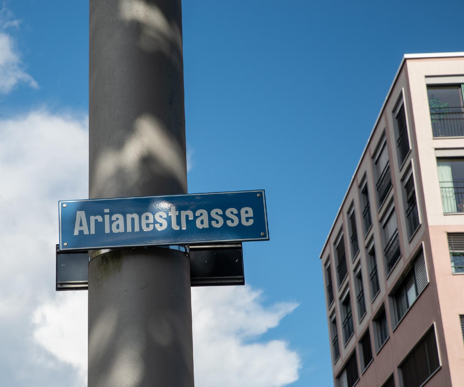 Blue street sign for Arianestrasse on a grey pole, with a pink/brown building in the background