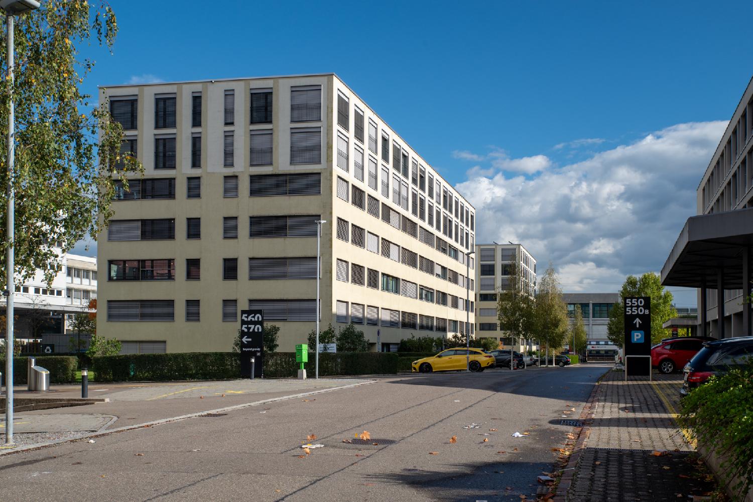 A street with 6-story office buildings and a few cars parked on parking lots. A train is passing in the background.