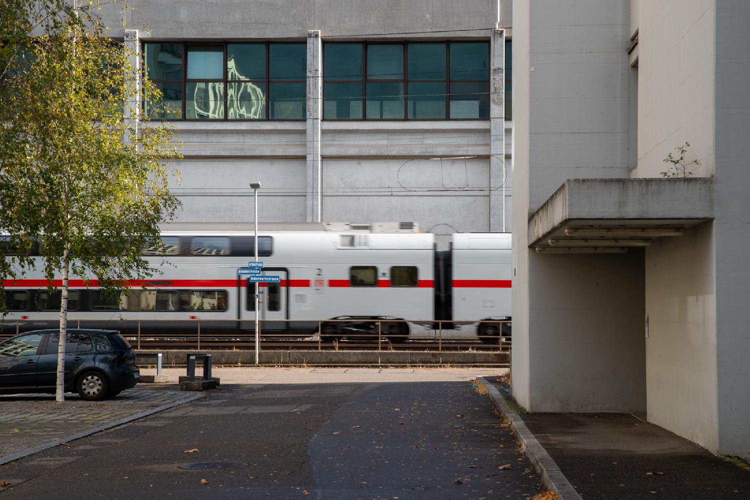 The end of a street, with a building in the background, and a white and red Deutsche Bahn ICE train moving on tracks perpendicularly to the street.