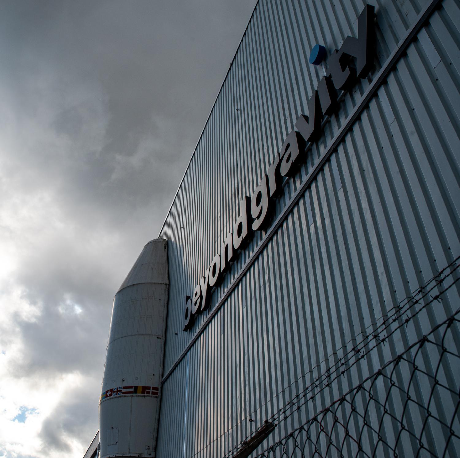 A metallic wall with half a rocket head on its side, and the Beyond Gravity logo on the wall. There's a metallic fence with barbed wire visible at the bottom of the picture.