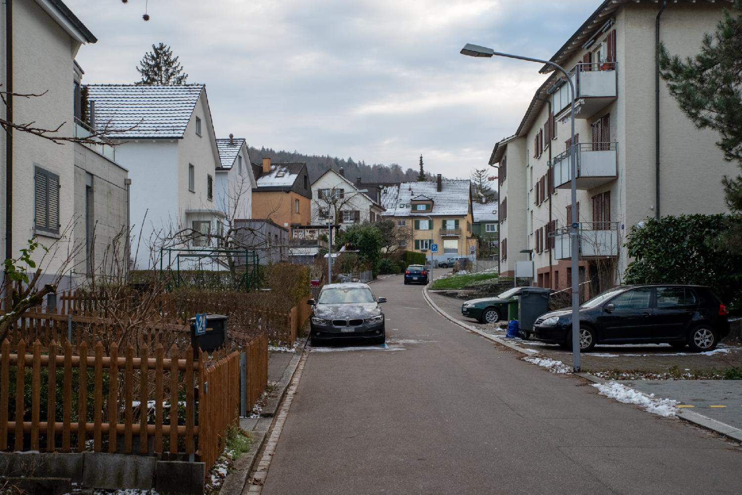 A narrow street with 3-4 story-buildings, some wooden fences and a few cars parked on the street and near the buildings. There is snow on the rooftops.