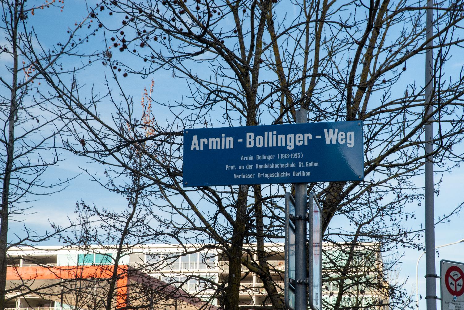 Blue "Armin-Bollinger-Weg" sign in front of trees and buildings. The sign reads: "Armin Bollinger (1913-1995). Prof. an der Handelshochschule St. Gallen. Verfasser Ortgeschichte Oerlikon".