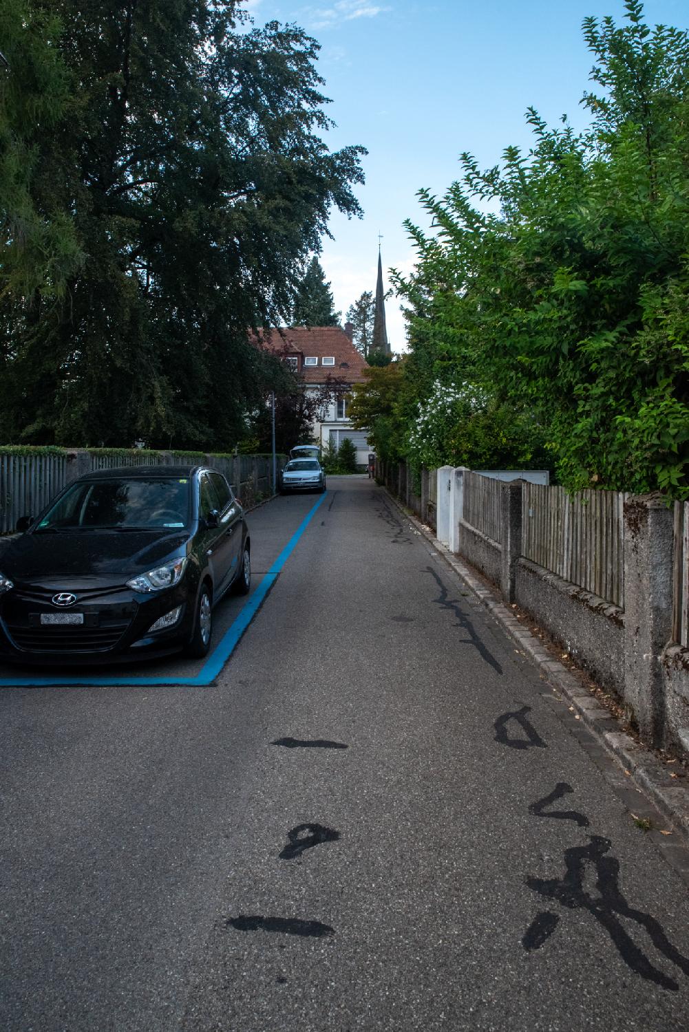 A narrow, one-way residential street with wooden fences on both sides, some cars parked in blue parking spots on the left, and a church tower in the background.