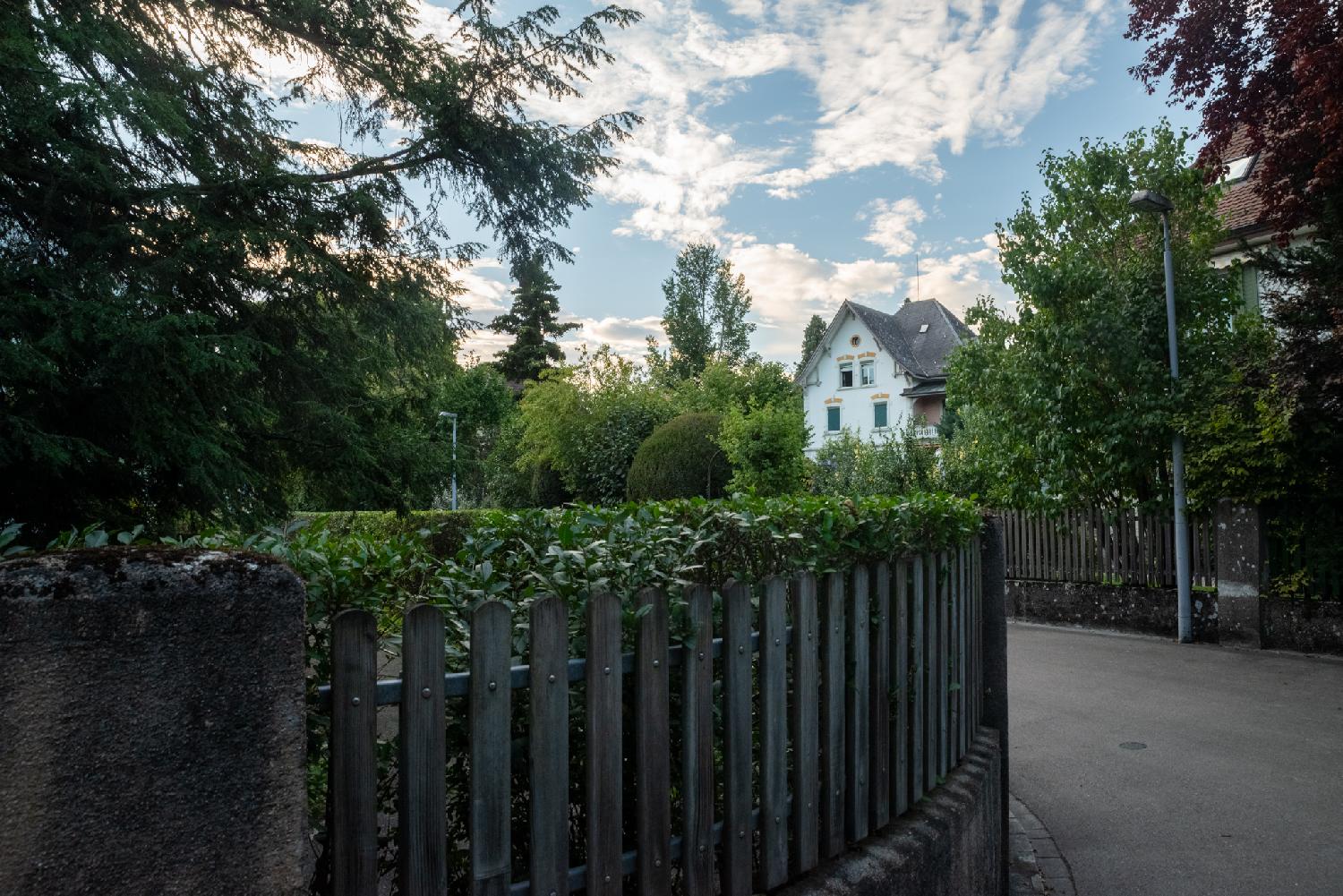 A curved residential street with wooden fences and greenery on each side, and a 3-story house in the background
