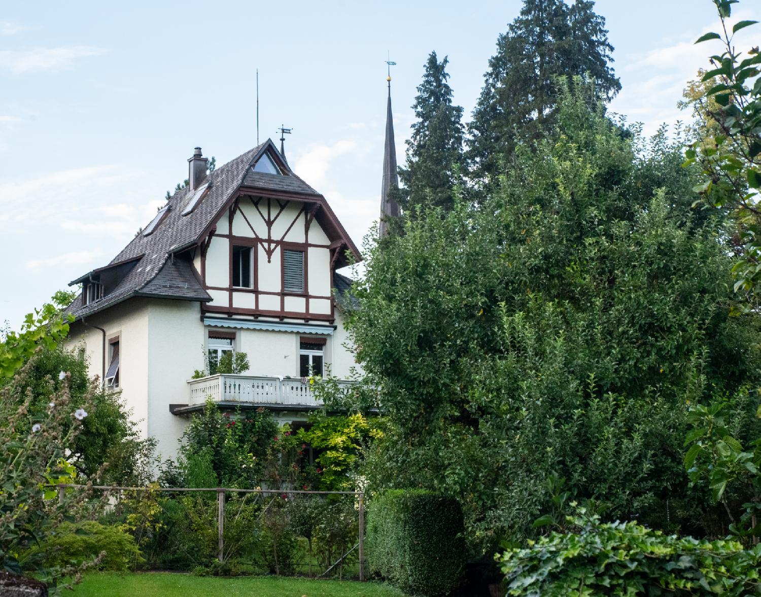 A half-timbered 3-story house in the middle of gardens with trees, hedges and flowers.