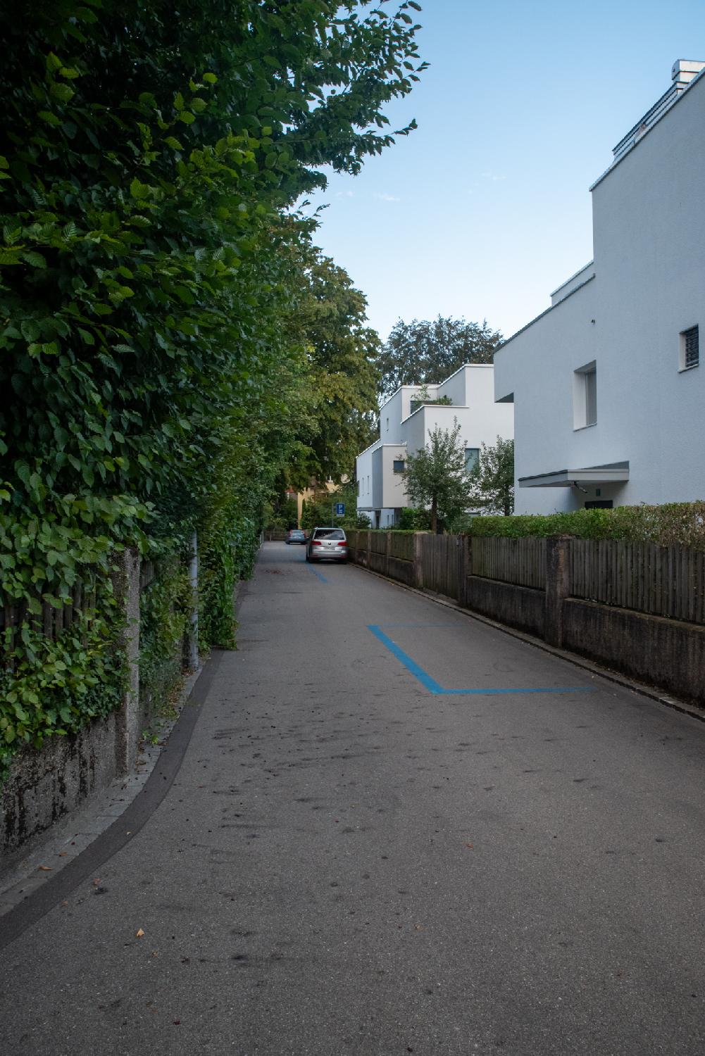 A narrow one-way residential street with greenery on the left and 3-story white habitation buildings on the right. There are blue parking spots and a couple of cars parked on the street.