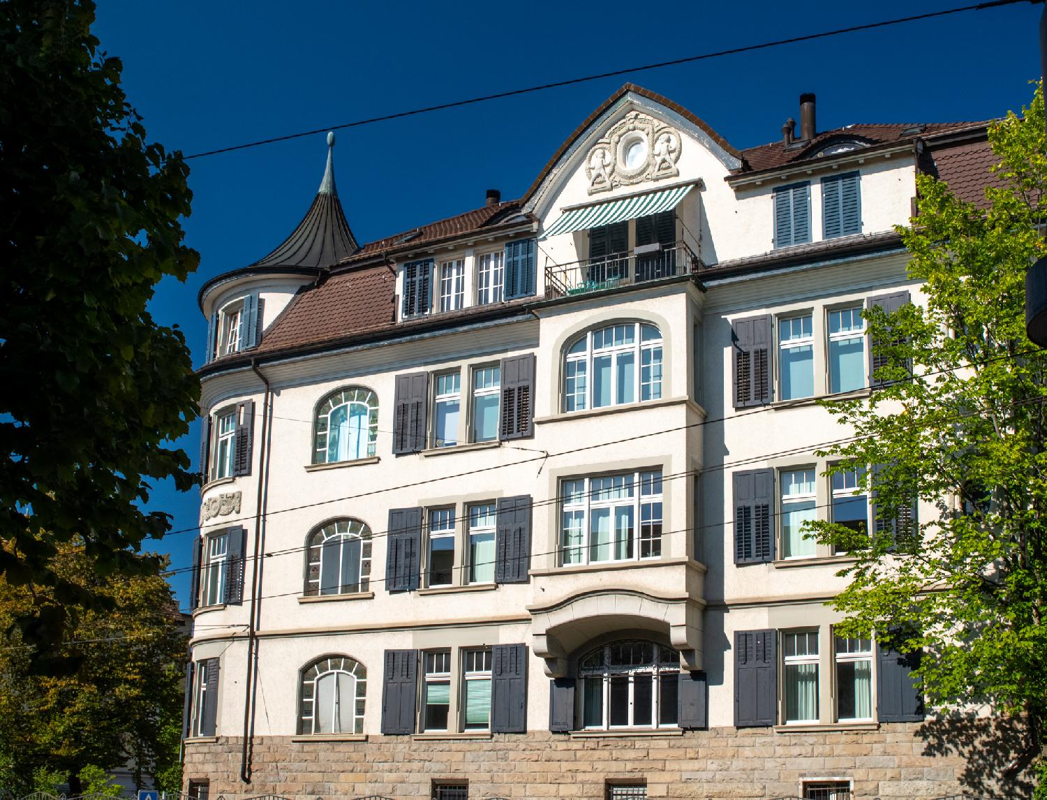 A four-story building. The corner is a small round tower with a concave conical roofs; the top of the building has moldings, and there are curved windows on the facade.