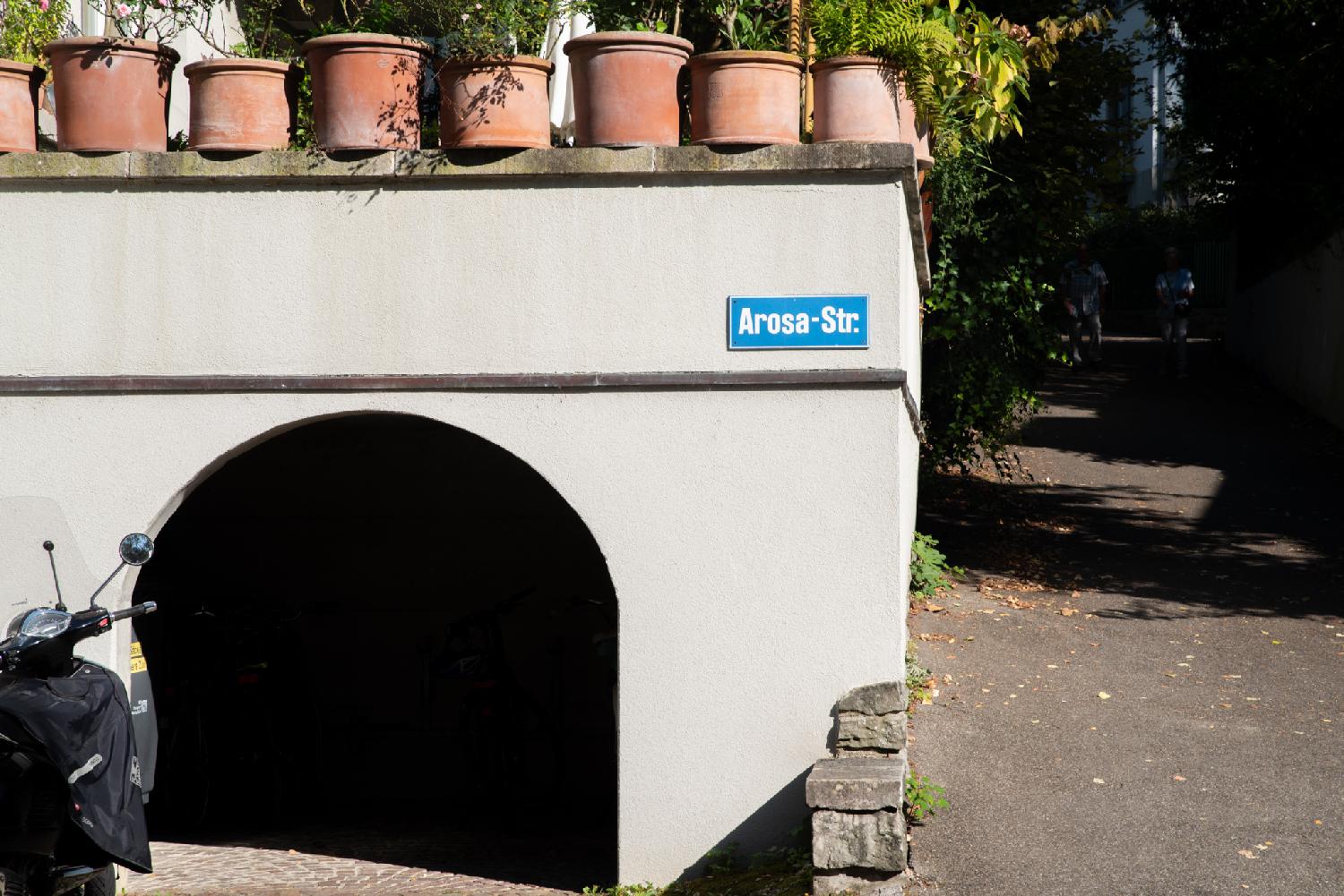 A blue "Arosa-Str" sign on a wall that has an arched opening and terracotta pots on top of it