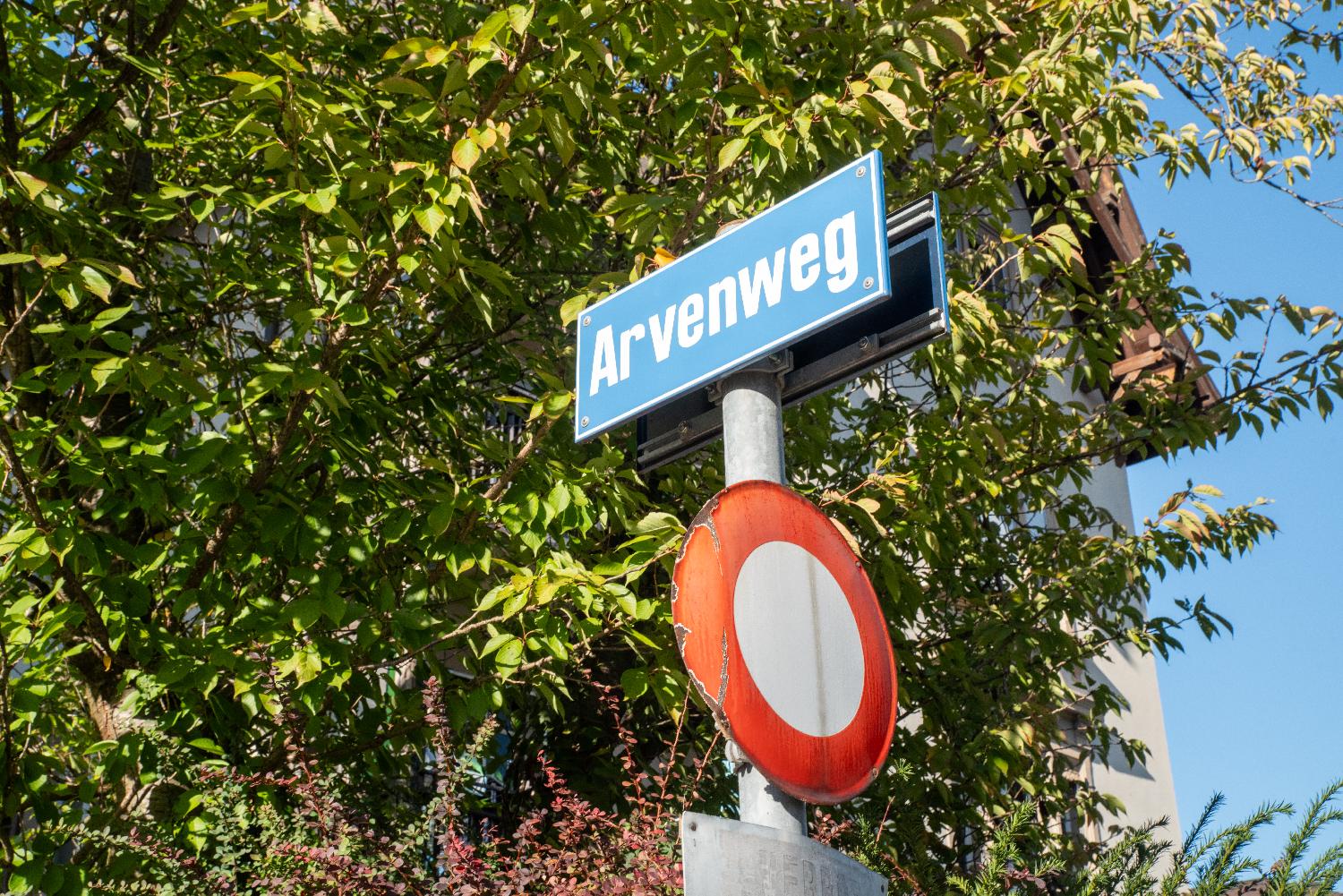 A blue "Arvenweg" sign in front of foliage, with a "road closed" white and red round sign below it