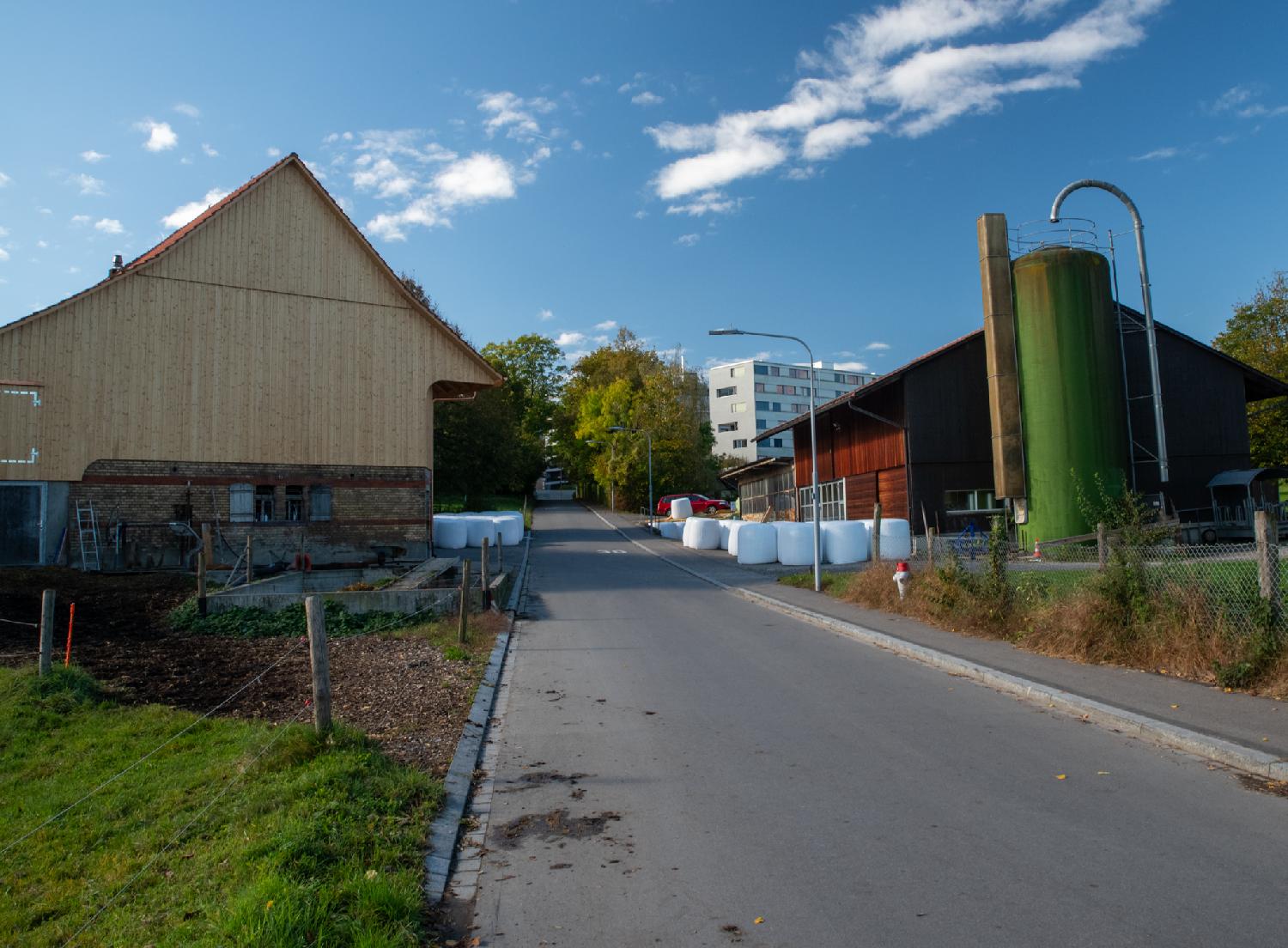 A road with farm buildings; on the left, a light-colored wooden building; on the right, a darker building with a green silo on the side. There are wrapped bales next to the buildings.