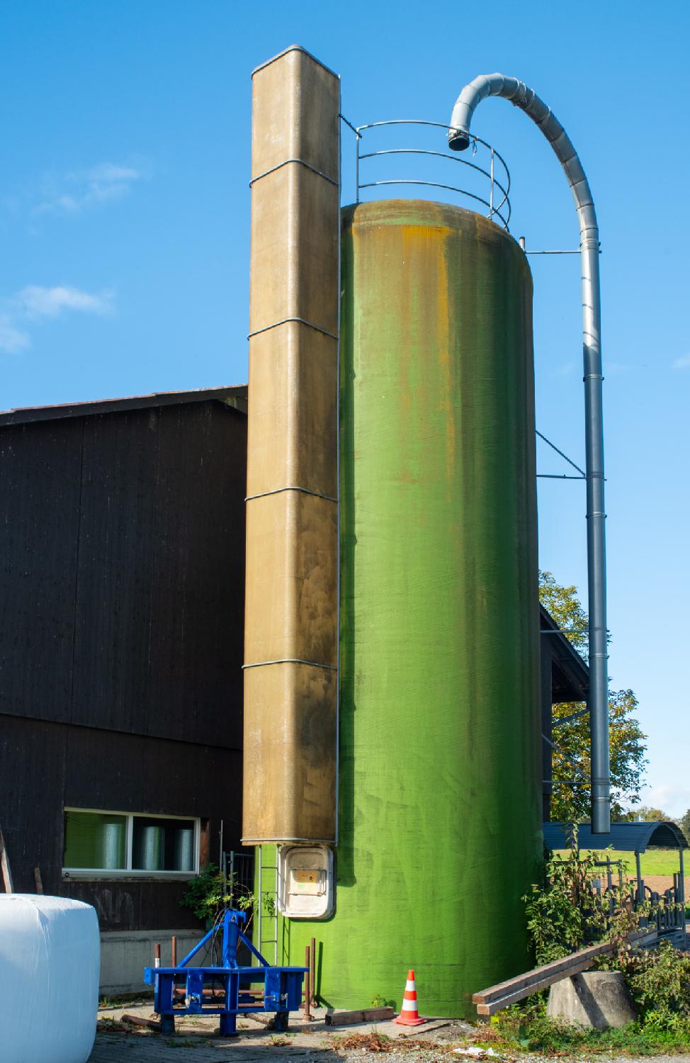 A green silo on the side of a building with a metallic curved pipe leading to it