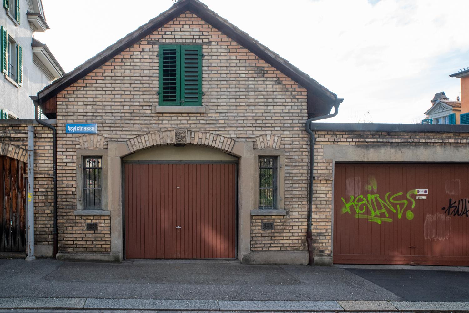 A small light brown brick building with two garage doors. A "Asylstrasse" blue street sign is attached to it.
