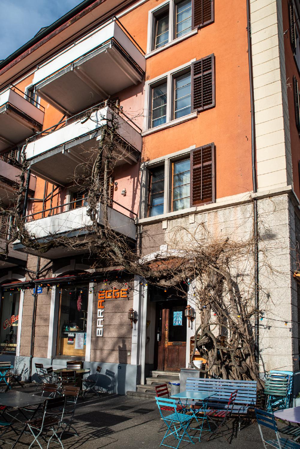 An orange residential building with a bar ("Bar Am Egge") on the ground floor, with an terrace and a gnarled tree climbing from the right side of the door up to the balconies on the right side of it