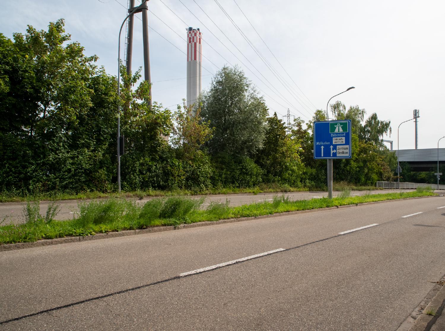 A 2x2 street with a sign to the highway and a high chimney (Aubrugg heat and power plant) with red and white checkered patterns on top in the background