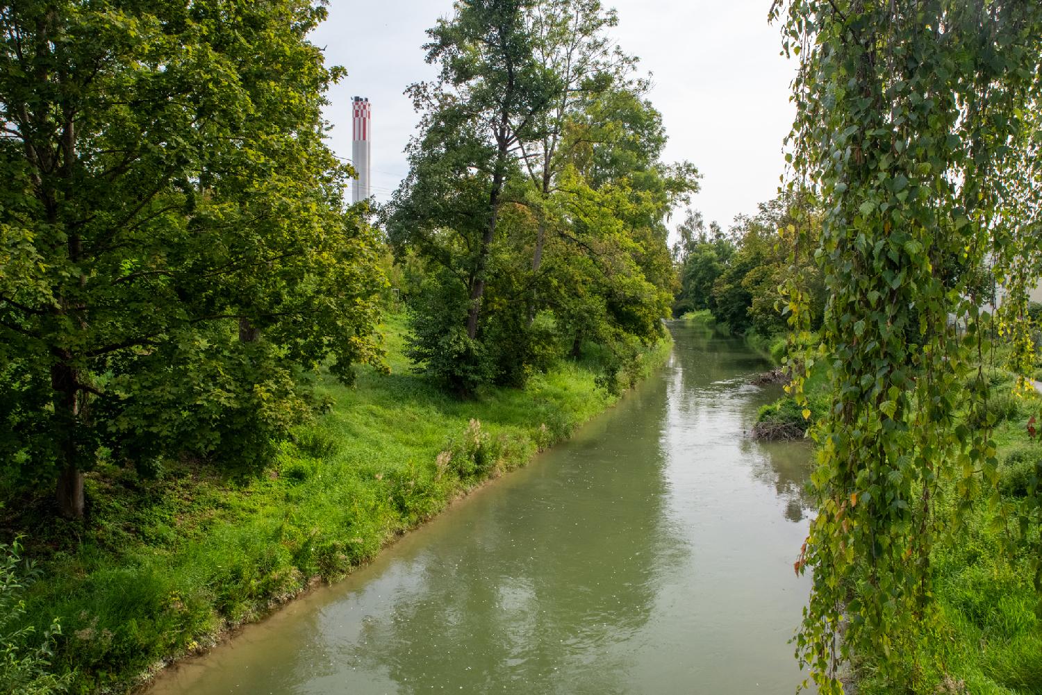 A narrow river with trees and grass on both banks. An industrial chimney is visible in the background.