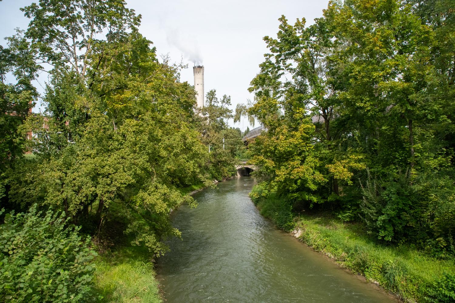 A narrow river with trees and grass on both banks, an small stone bridge further along the river. An industrial chimney is visible in the background.