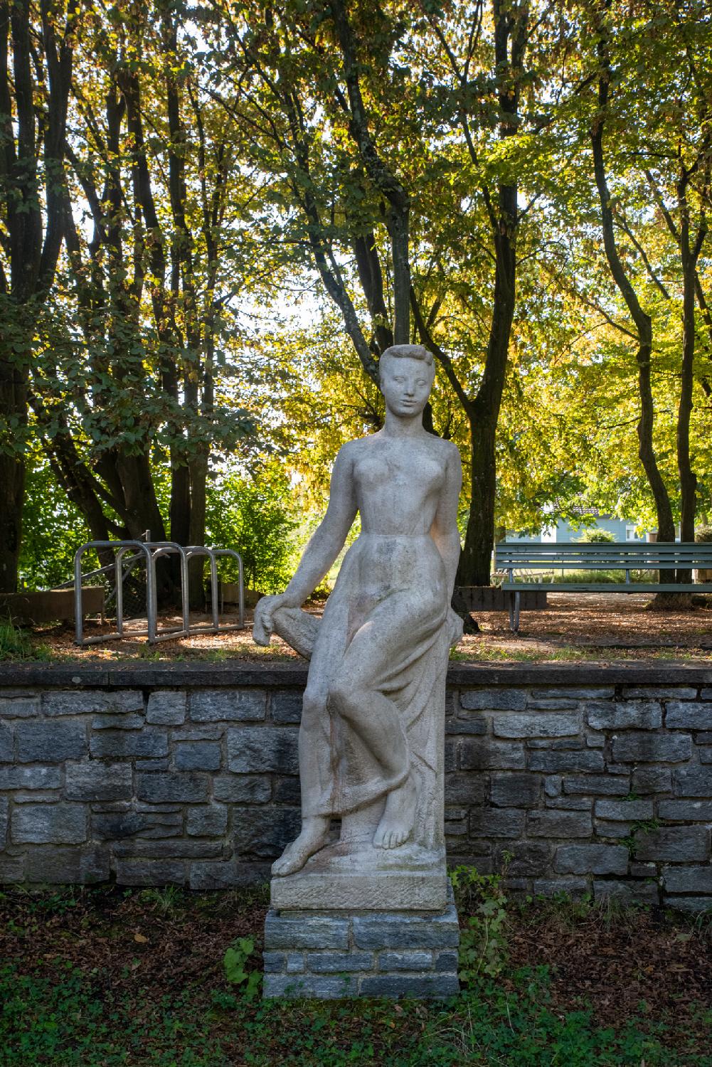 White statue of a woman leaning/sitting on a high chair, in a park with trees and a bench behind it