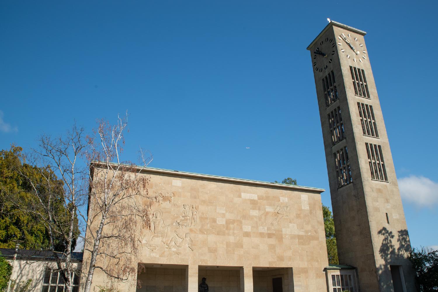A church building composed of a light brown parallelepipedic building and a church tower with narrow openings and clocks on each side.