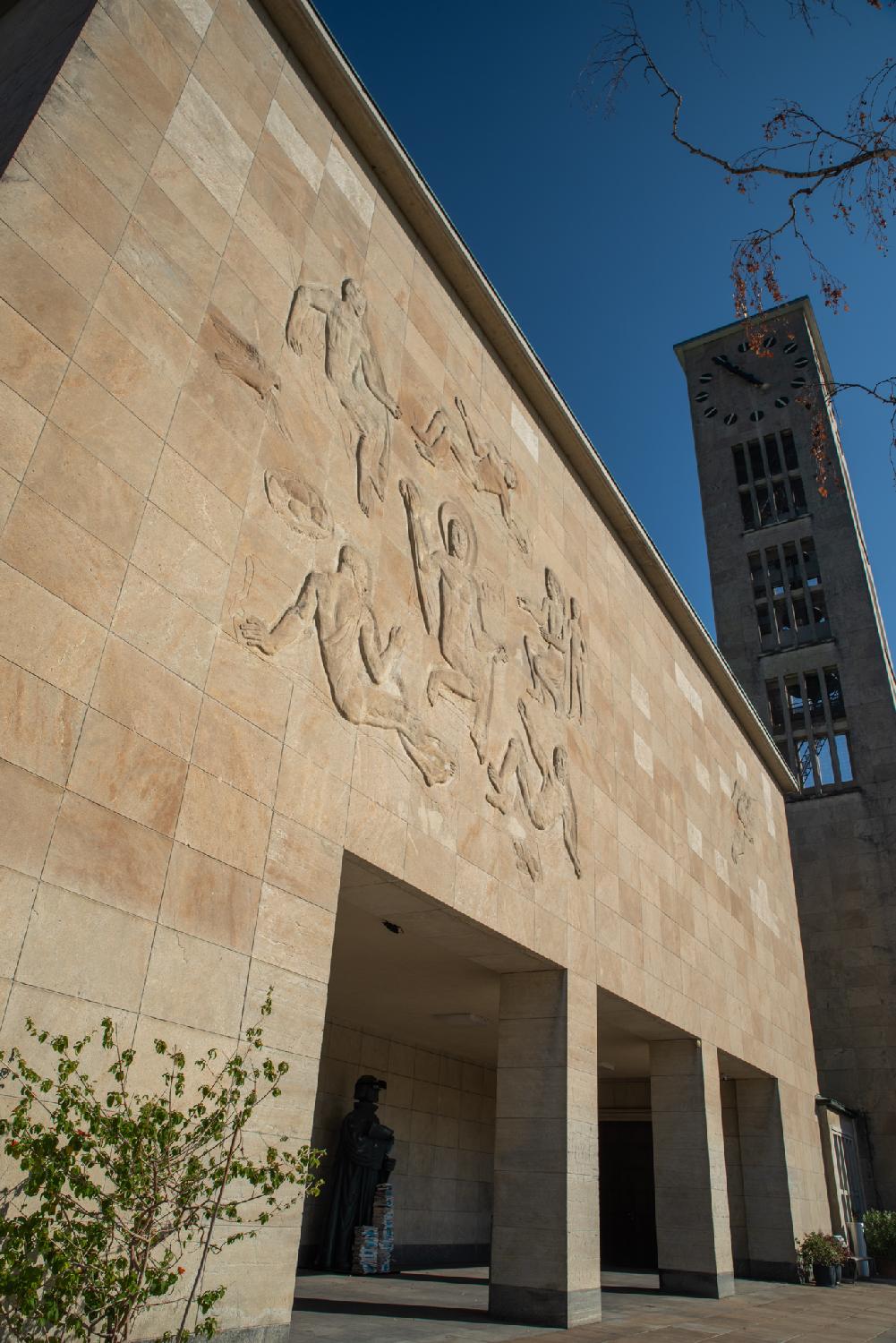 A brown wall with reliefs depicting Feeding the multitude (mural by Otto Bänninger), with a church tower in the background.