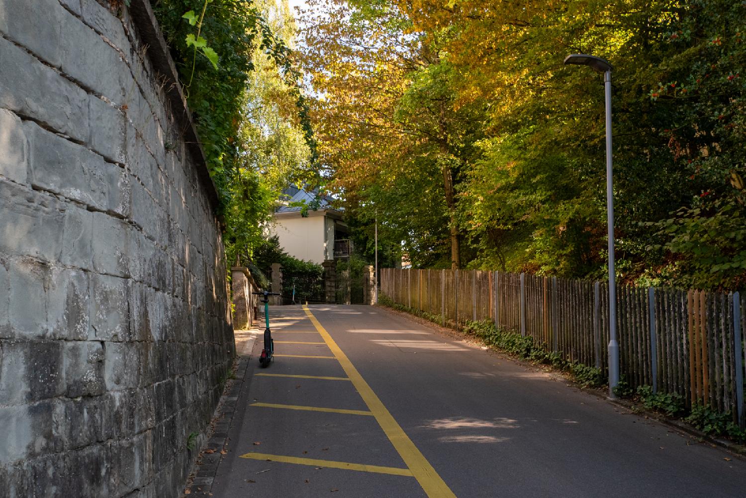 A narrow street with painted yellow zebras, a stone wall on the left and a wooden fence on the right with trees behind it. There's an electric scooter parked on the zebras.