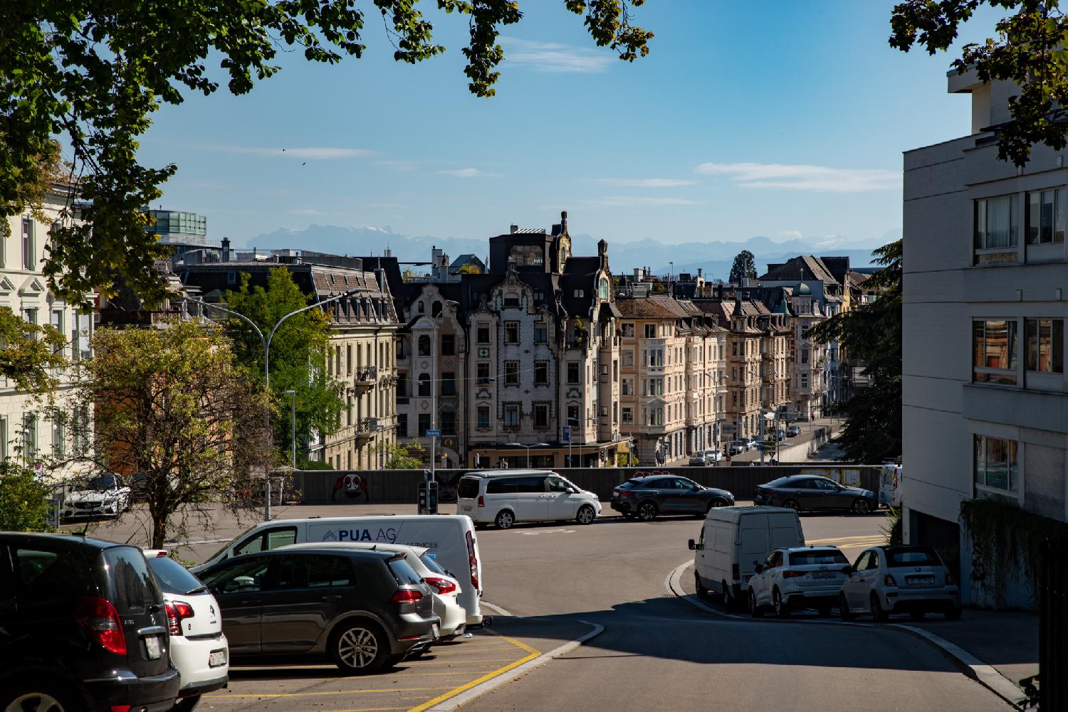A street going down, with cars parked on each side and on the perpendicular road. In the middleground, some buildings on a lower-level street. In the far background, the Alps, quite hazy.