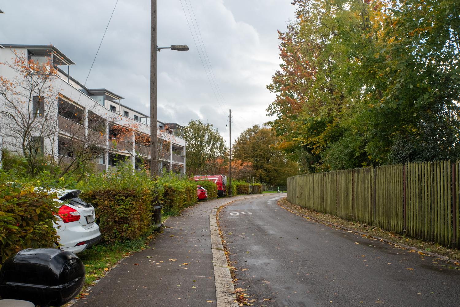 A narrow road with 3-storey residential buildings on the left and a wooden fence with trees on the right.
