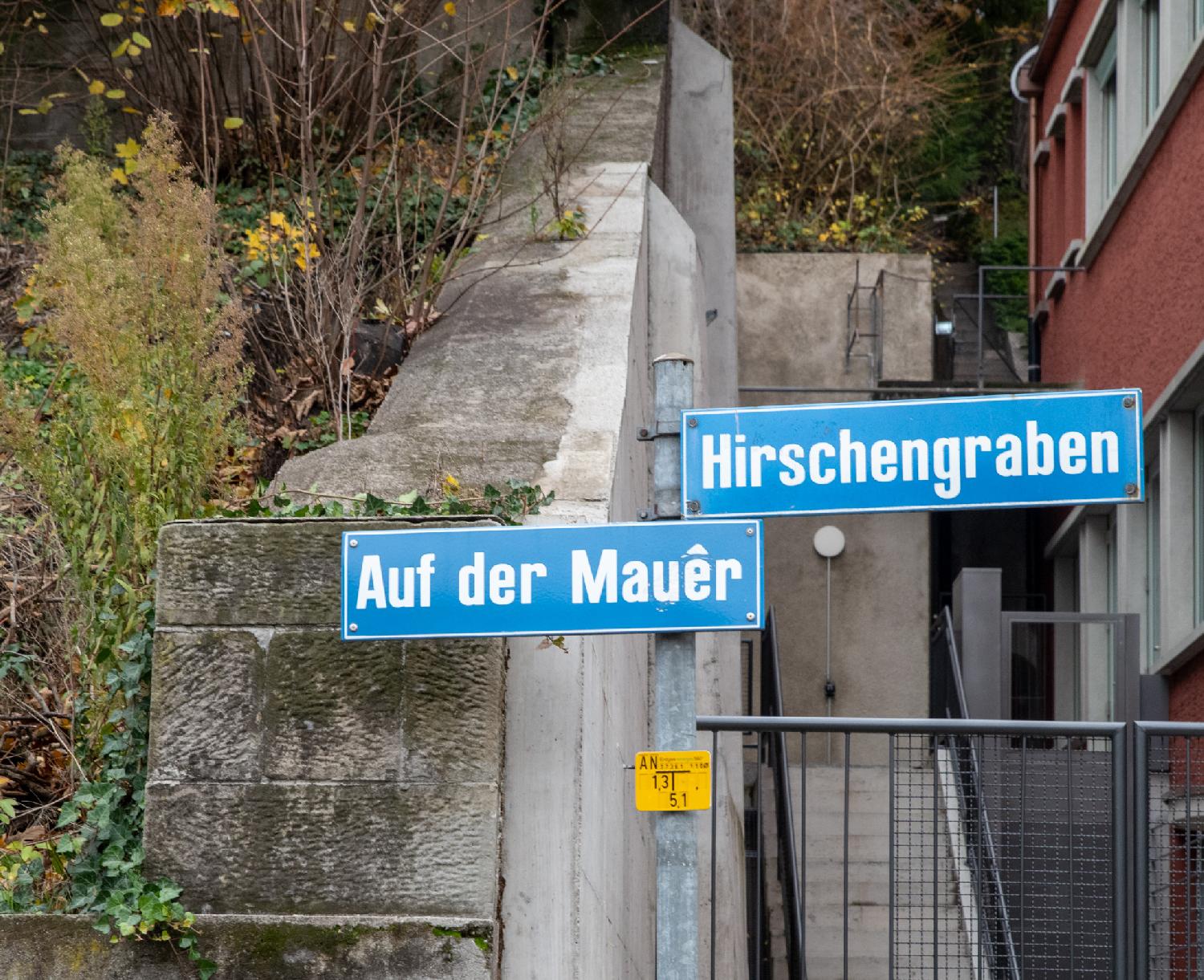 Two blue street signs for Auf der Mauer and Hirschengraben in front of a building, a separating wall and some vegetation.