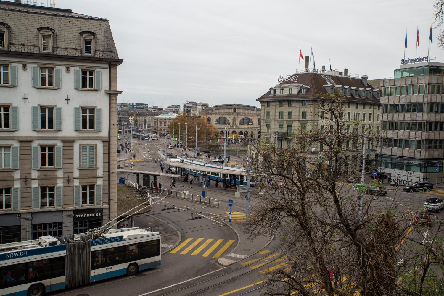 A large street intersection with a tram stop in the middle.There are a tram and a trolleybus visible on the picture, as well as buildings including the main station of Zürich.