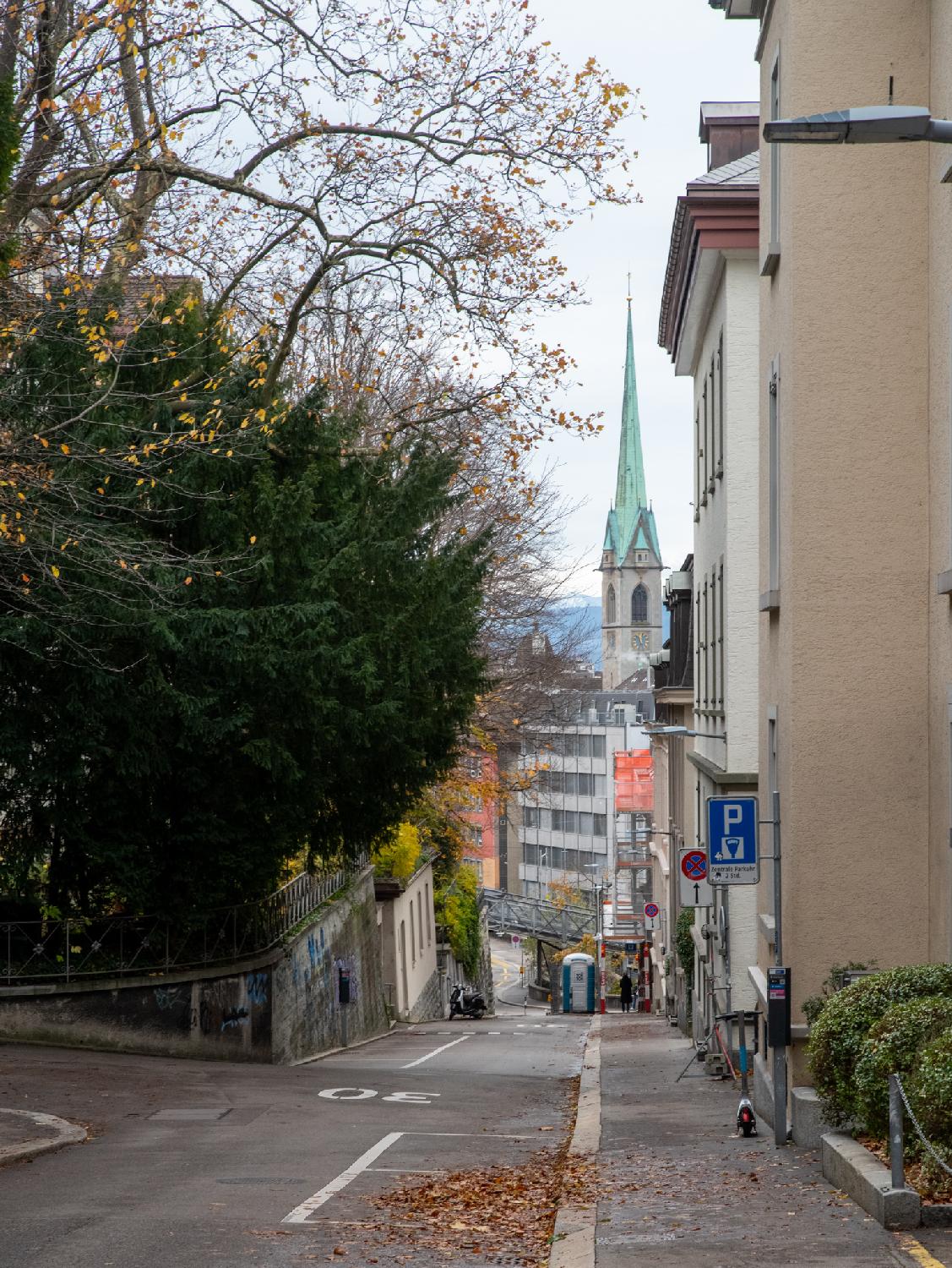 A street going downhill, with buildings on the right and trees on the left, with a church tower with a green steeple in the background
