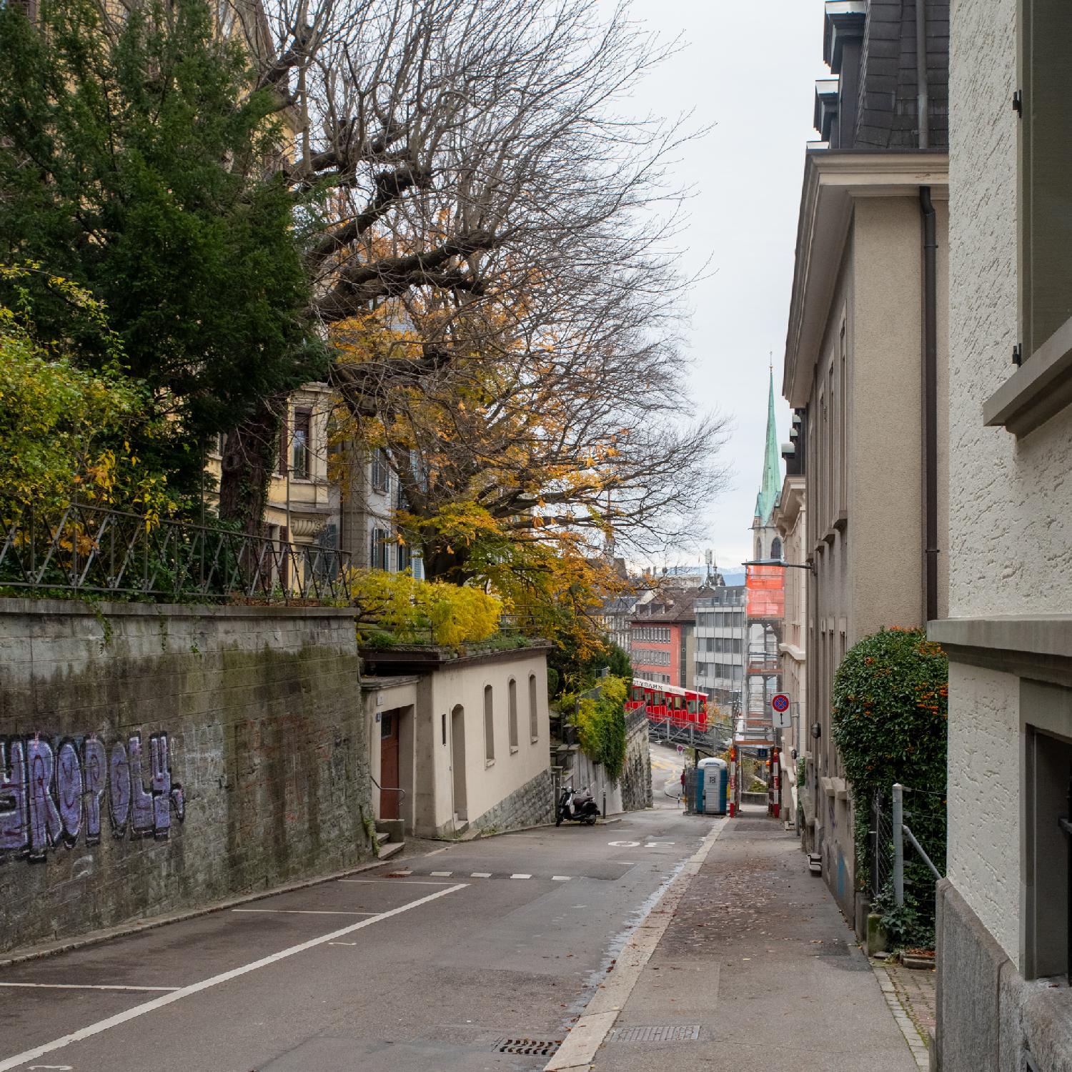 A street with a wall and a small building, with larger buildings on top, on the left, and buildings on the right; in the background, a red funicular going downhill.