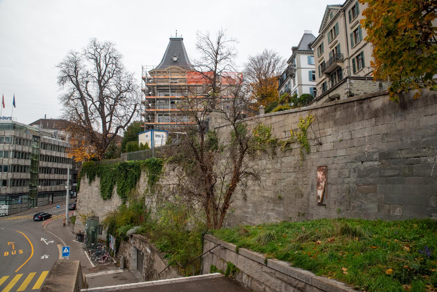A wall next to a road, on top of which there is a street with buildings. The wall is covered in ivy and other vegetation.