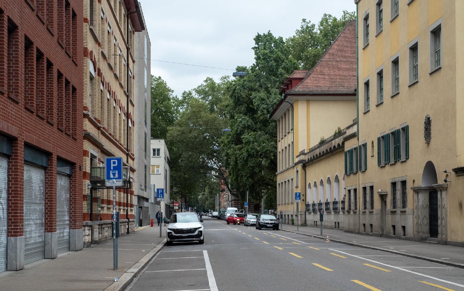 A city street with 4-5-story buildings and cars parked on each side. There are trees in the back of the street/background of the image.