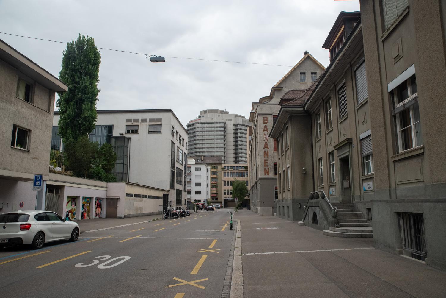 A street with 3-4-storey buildings in grey/brown shades, with a higher building visible at the end of the street. One of the buildings has "Bananen" written on the side of it.