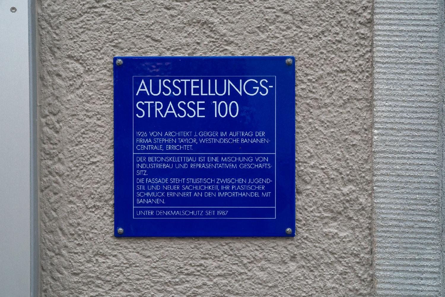A blue sign on a concrete wall, reading the following German text: "Built in 1926 by architect J. Geiger on behalf of the Stephen Taylor, Westindische Bananen-Centrale. This concrete skeleton frame building is a blend between an industrial building and business headquarters. The façade's style stands between Art Nouveau and New Objectivity; its sculptural decorative elements remind of the banana import trade. Protected building since 1987."