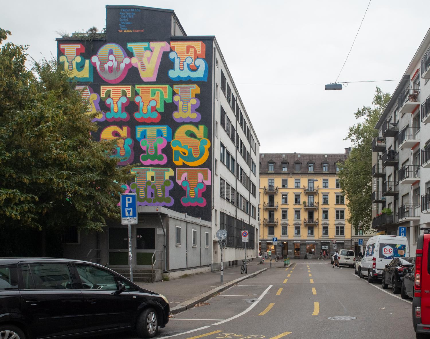 A street with 5- storey buildings on each side. The side of the left building is painted with a colorful lettered mural reading Love At First Sight
