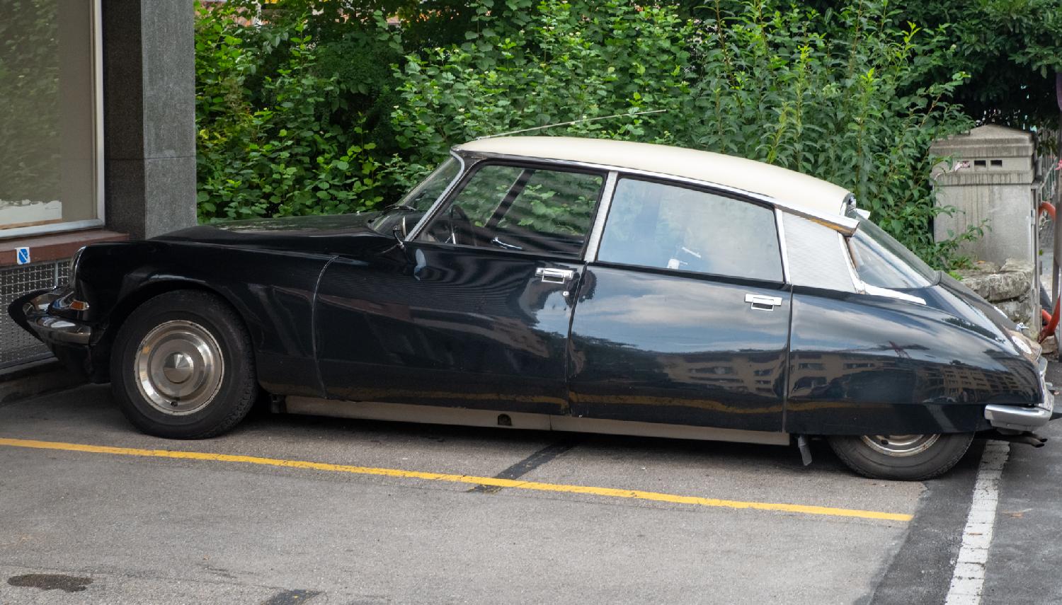 A black and white Citroen DS, seen from the side, parked in front of a building and a hedge