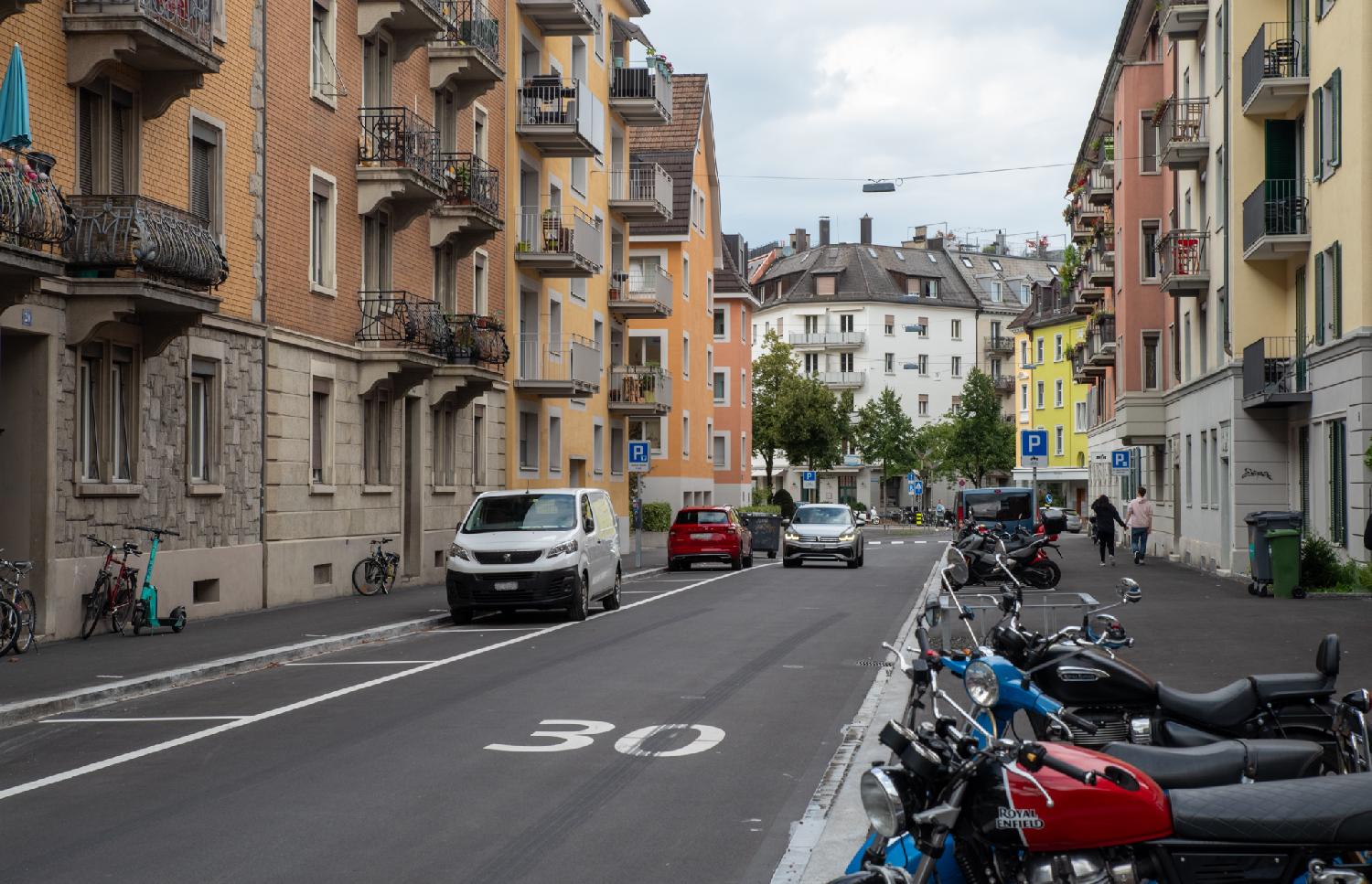 A street with a 30km/h speed limit painted on the road, with 3-4 storey-buildings on each side, some car parking spaces on the left and a few motorbikes parked on the right