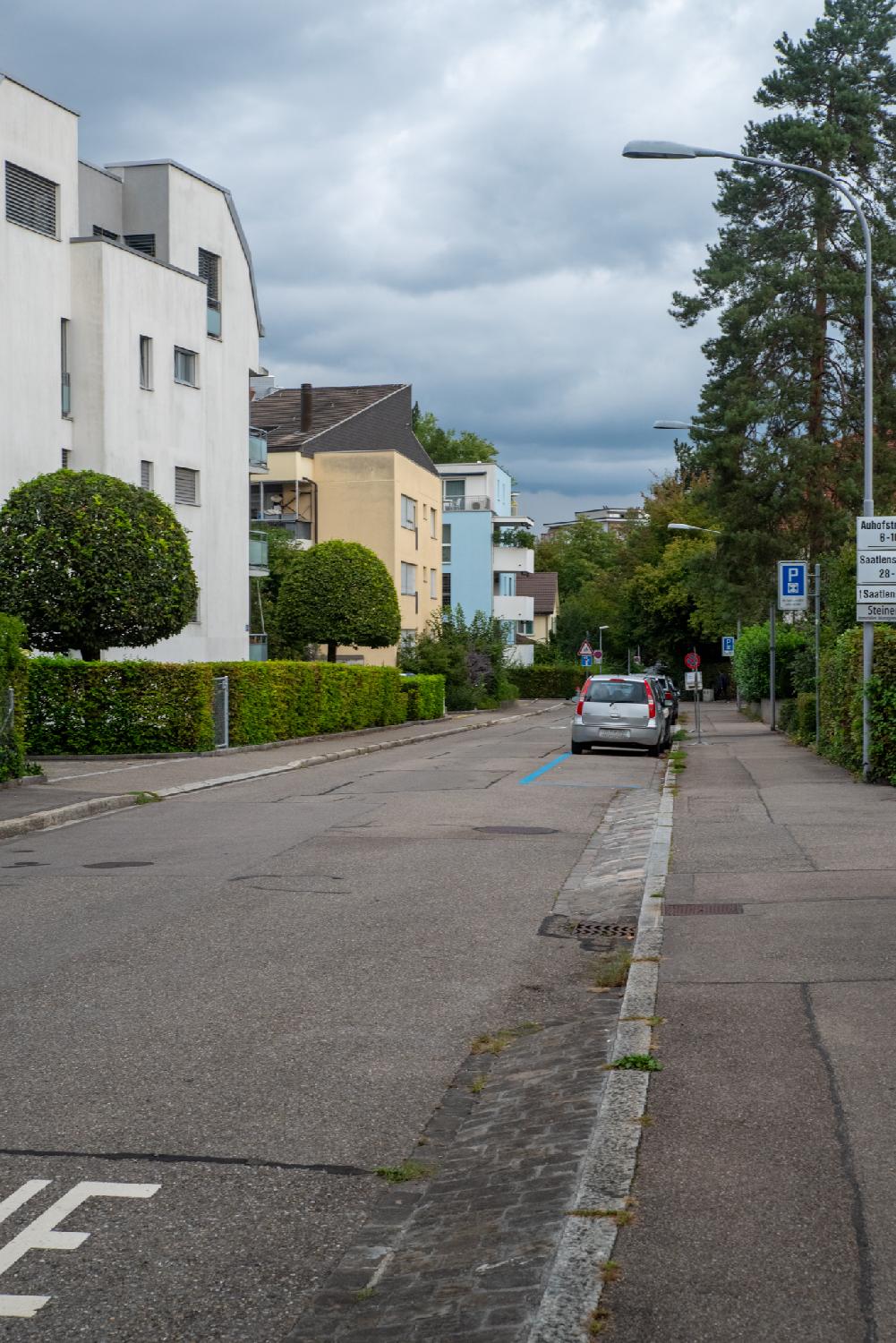 A residential street with 4-5-storey buildings on the left side, hedges and trees with cars parked on the side of the street on the right side.