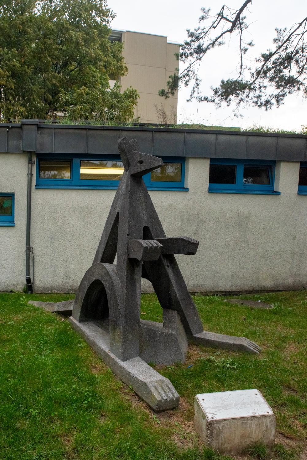 A stylized sculpture of an animal (a kangaroo?) in front of a low building with small, high windows.
