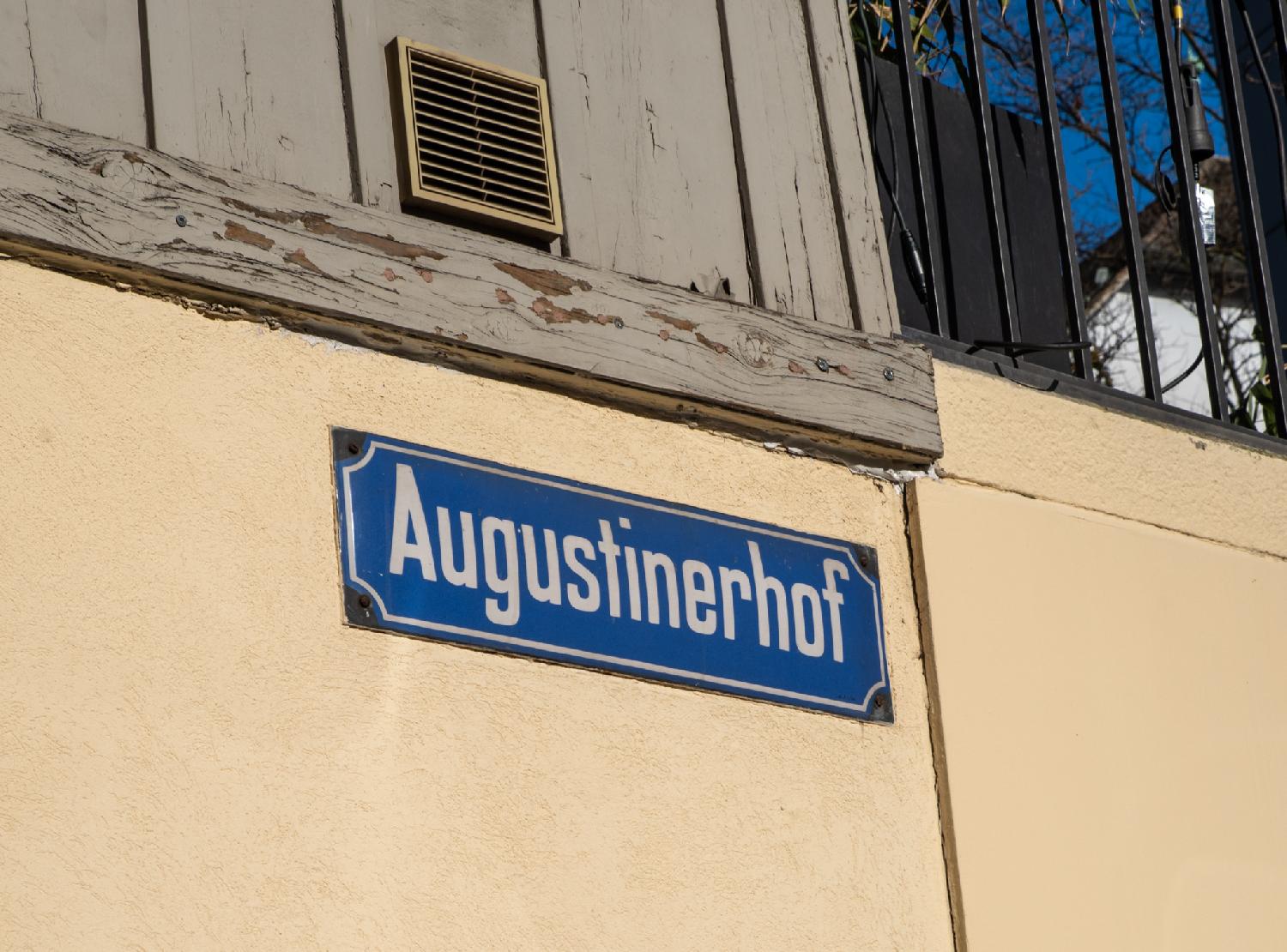 A blue "Augustinerhof" sign on a yellow concrete wall below a wooden wall part