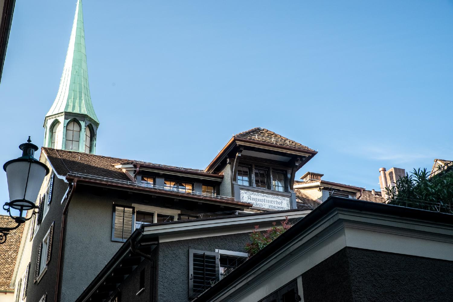 Rooftops at various heights, with a "Augustinerhof" sign at the bottom of a 3-pane dormer window. A green church steeple is visible in the background.