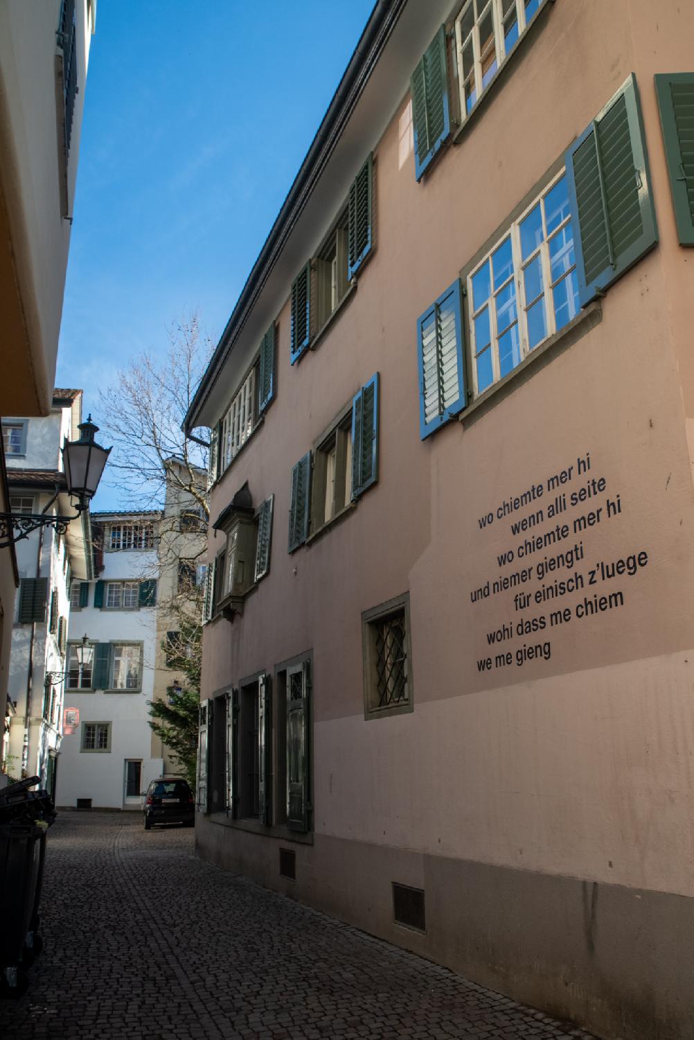 A narrow alley with a 3-storey building with green blinds, with a poem written on the wall: wo chiemte mer hi wenn alli seite wo chiemte mer hi und niemer giengti für einisch z'luege wohi dass me chiem we me gieng