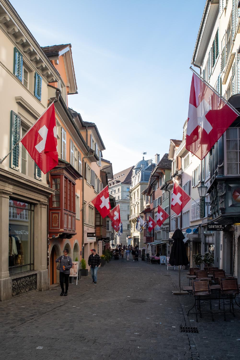 A pedestrian alley with 4-storey buildings on each side, shop fronts on the ground floor, and a lot of Swiss flags hanging from the buildings