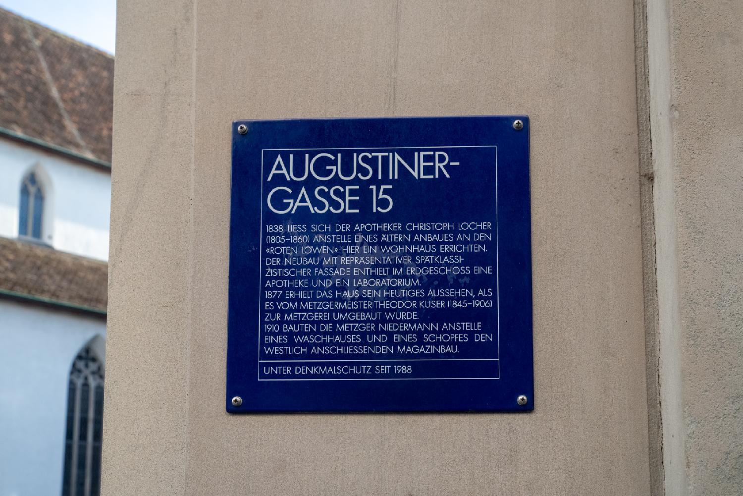A blue sign on a wall, with a church-like looking building in the background. The sign reads "Augustinergasse 15 1838 liess sich der Apotheker Christoph Locher (1805-1860) anstelle eines ältern Anbaues an den "Roten Löwen" hier ein Wohnhaus errichten. Der Neubau mit repräsentativer spätklassizistischer fassade enthielt im Erdgeschoss eine Apotheke und ein Laboratorium. 1877 erhielt das Haus sein heutiges Aussehen, als es vom Mezgermeister Theodor Kuser (1845-1906) zur Metzgerei umgebaut wurde. 1910 bauten die Metzger Niedermann anstelle eines Waschhauses und eines Schopfes den westlich anschlissenden Magazinbau. Unter Denkmalschutz seit 1988."