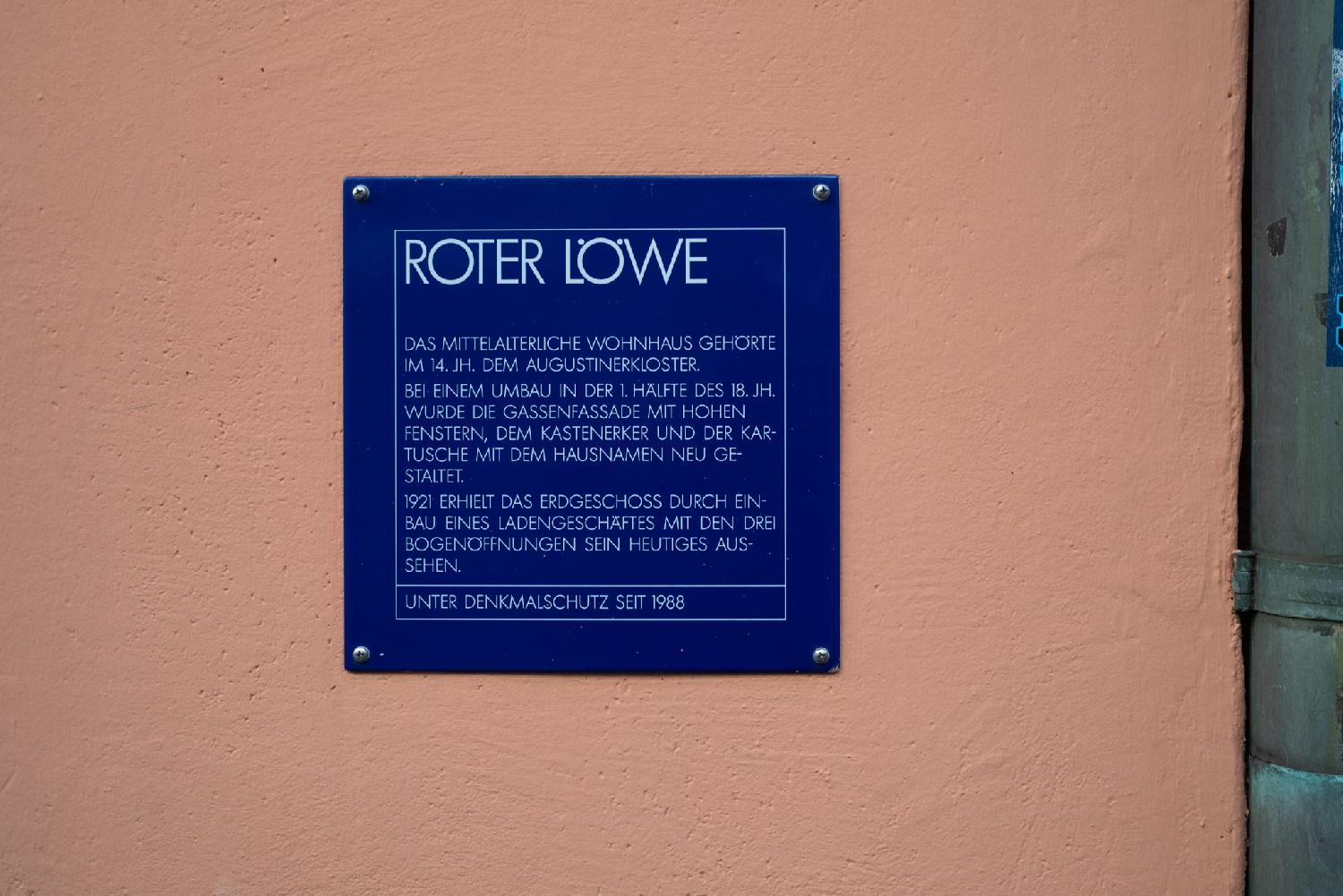 A blue sign on a coral-colored wall, reading: Roter Löwe Das mittlealteriche Wohnhaus gehörte im 14. Jh. dem Augustinerkloster. Bei einem Umbau in der 1. Hälfte des 18. JH. wurde di Gassenfassade mit hohen Fenstern, dem Kastenerker und der Kartusche mit dem Hausnamen neu gestaltet. 1921 erhielt das Erdgeschoss durch Einbau eines Ladengeschäftes mit den drei Bogenöffnungen sein heutiges Aussehen. Unter Denkmalschutz seit 1988