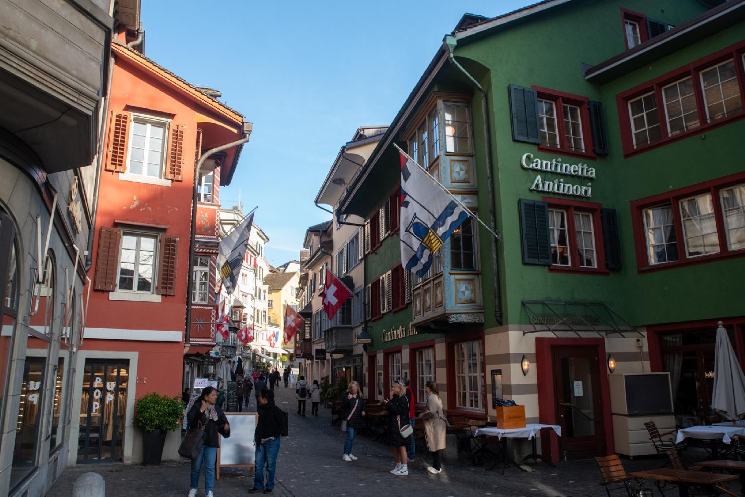 A pedestrian alleyway with shops and restaurants, 3-4 storey-buildings, a few oriel windows, and a dozen pedestrians.