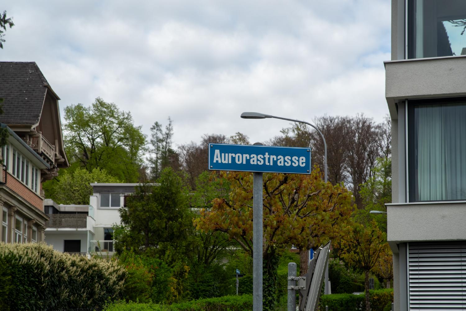 A blue "Aurorastrasse" street sign in front of gardens and houses