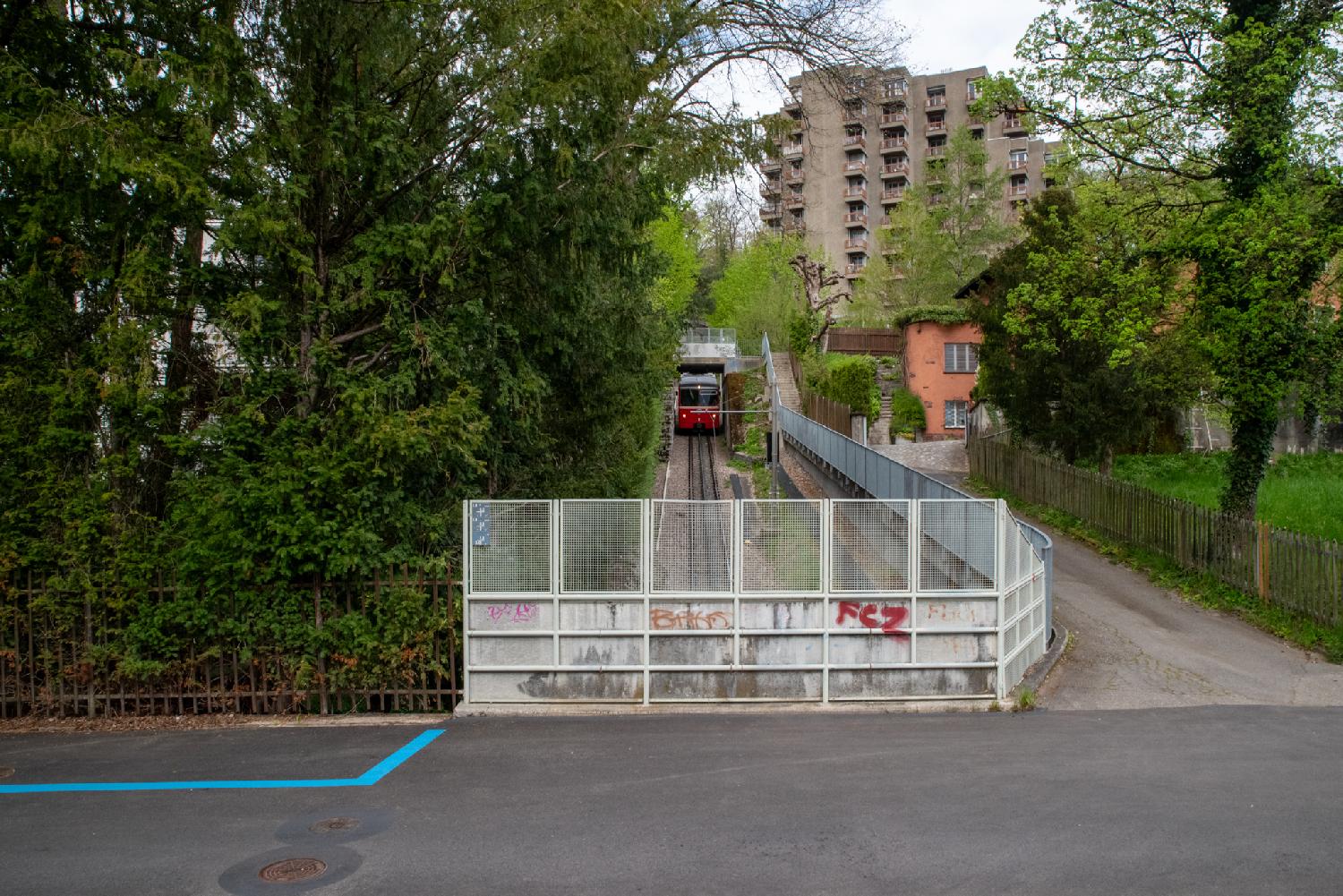A small red rack railway train, seen from a bridge above the railway, with trees and a large brown building framing the tracks.