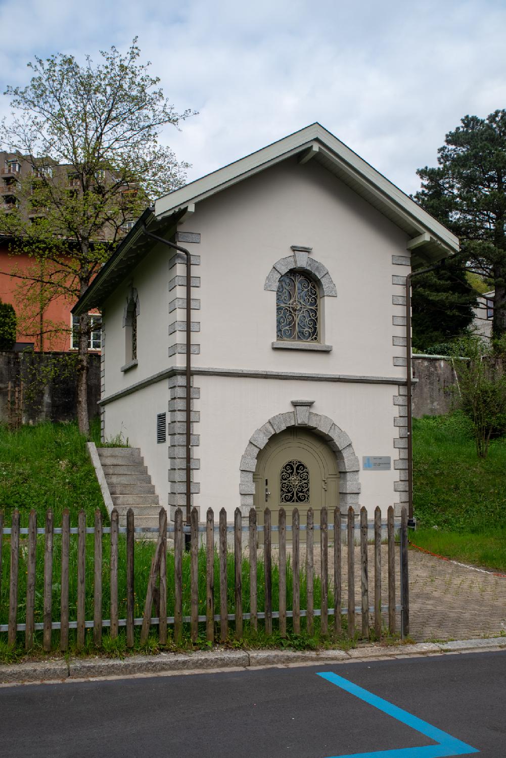 A small, two-storey white building with arched windows and doors and a staircase on the left side